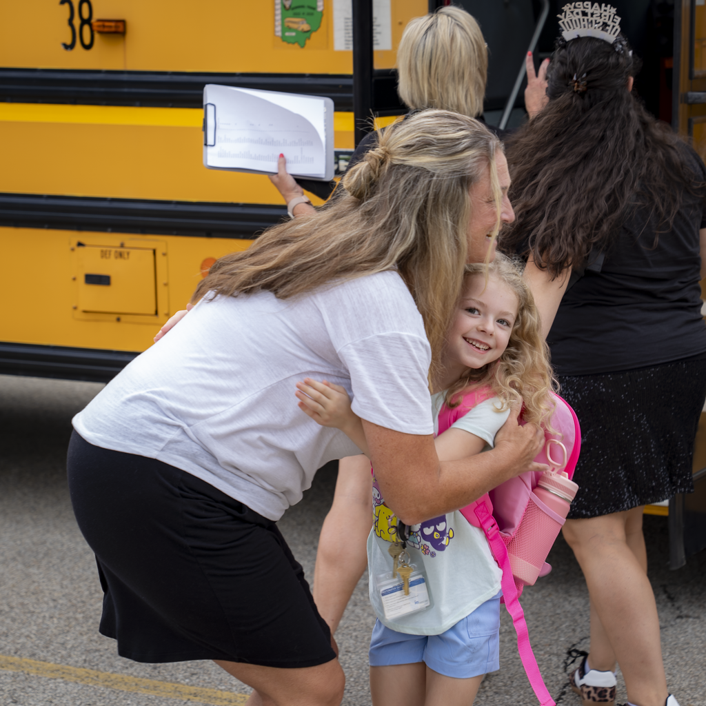 Girl getting a hug from a teacher in front of a yellow school bus