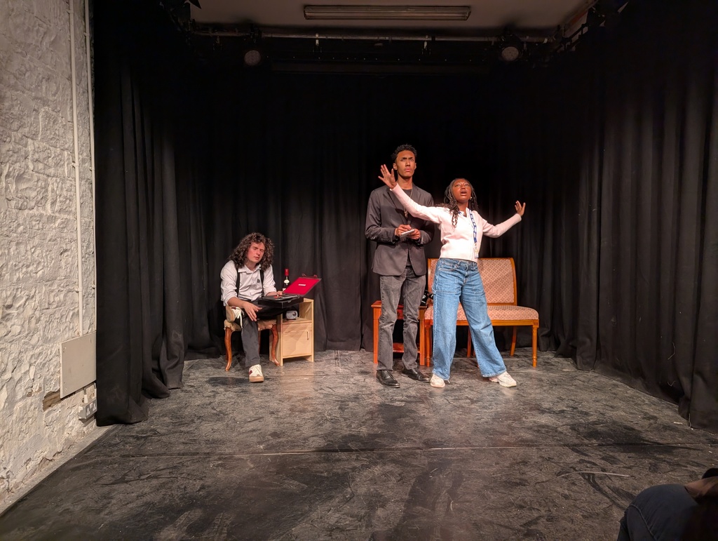 Man with curly hair at a table in the back next to another tall man behind a woman spreading her arms all on a stage