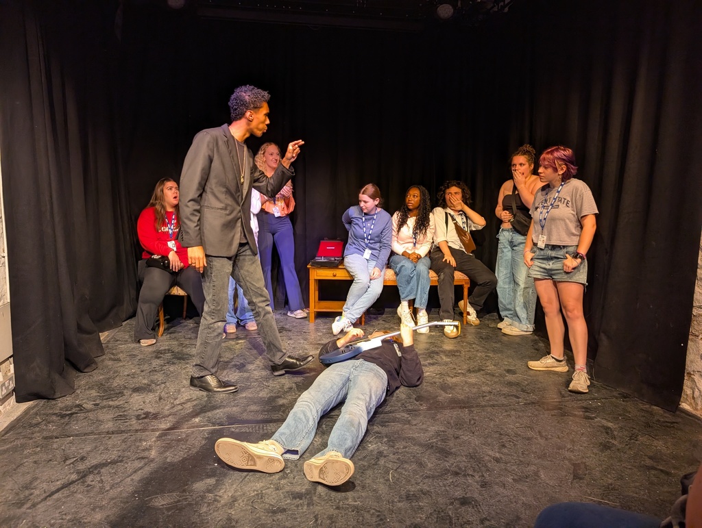 A tall man in a black suit pointing a teens on a stage while a person lies on the floor with a guitar in hand