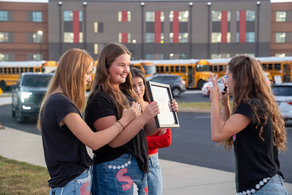 three girls holding a picture frame smiling at another girl on the opposite side of the frame