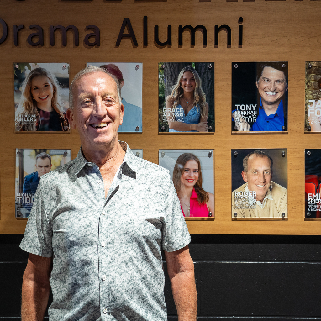 Older man in button down shirt smiling while standing in front of a younger photo of himself that is hung on a wall of Drama Alumni