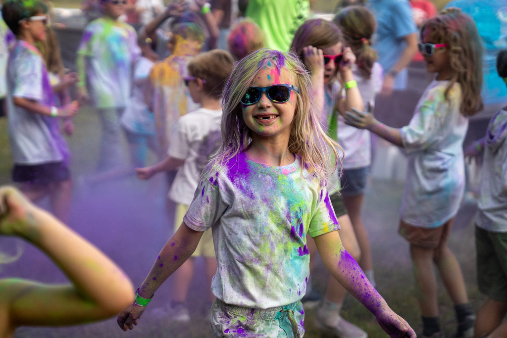 A student covered in paint wearing sun glasses and missing two front teeth smiling at the camera. 