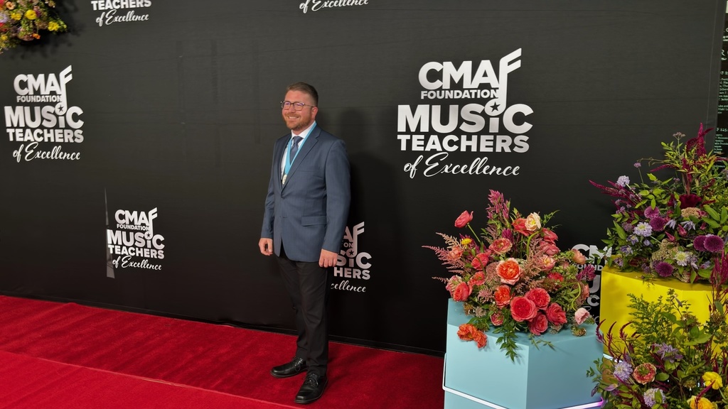 A man in a blue suit standing on the red carpet next to flowers in front of a banner saying "CMAF Foundation - Music Teachers of Excellence" 