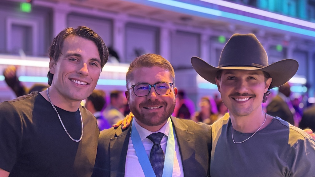 A man with glasses and a tie in between a taller man with a silver chain and a man with a mustache and a cowboy hat