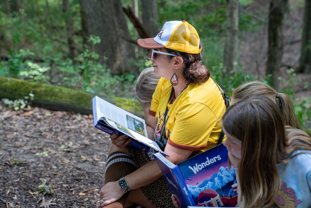 A woman in yellow with a yellow hat reading off a textbook smiling in the woods