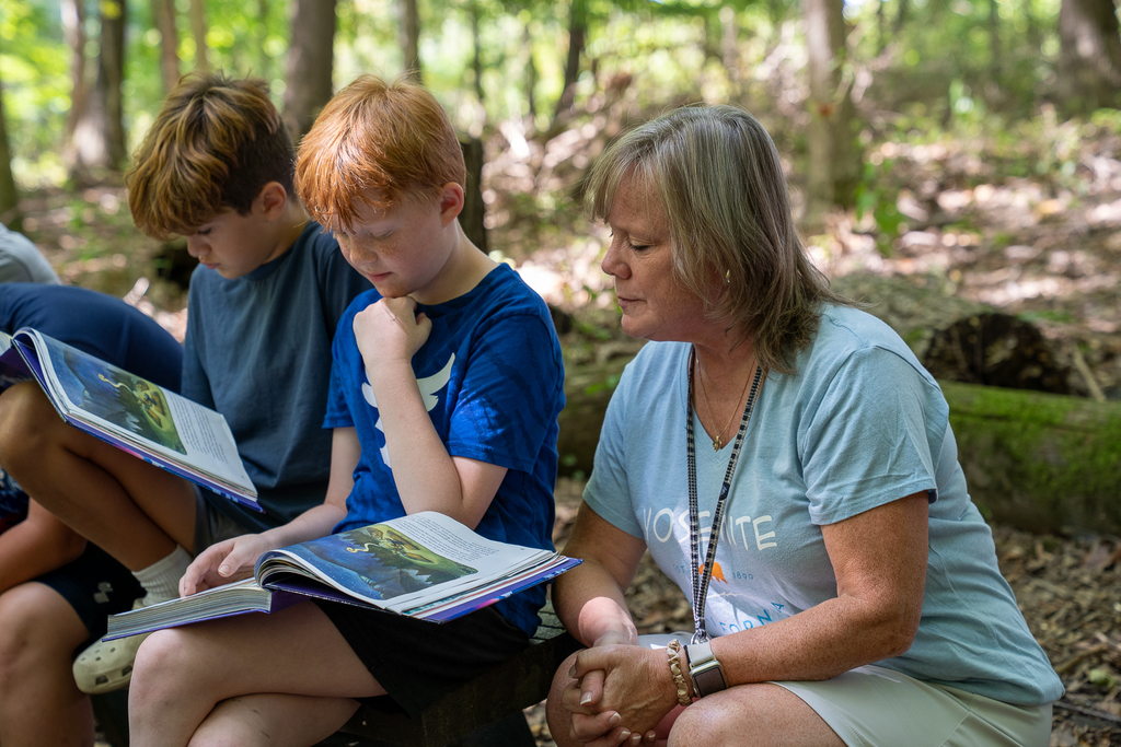 A teacher squatting down in the woods looking over the shoulder of a boy with red hair reading off a textbook