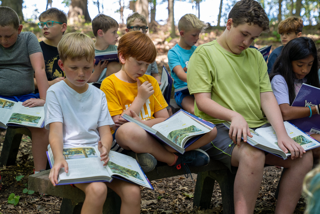 A group of boys reading in the woods while the boy in the middle has a yellow shirt and red hair reading criss cross
