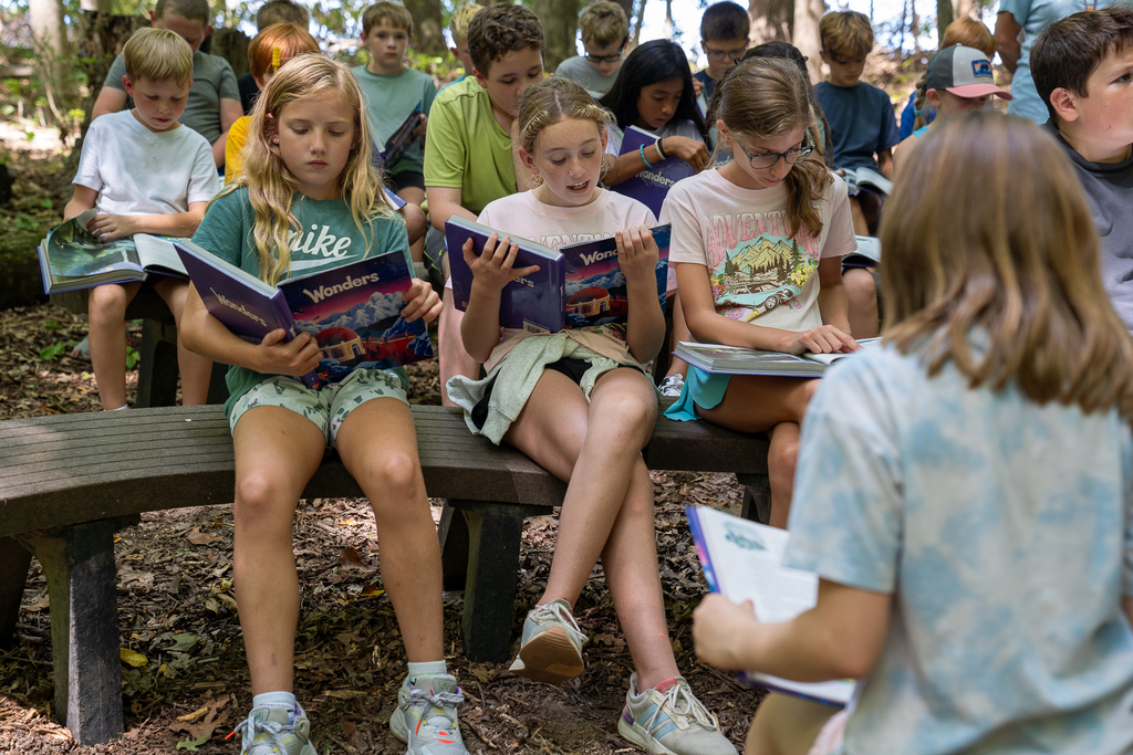 three girls on a bench in the middle of the woods reading from a textbook