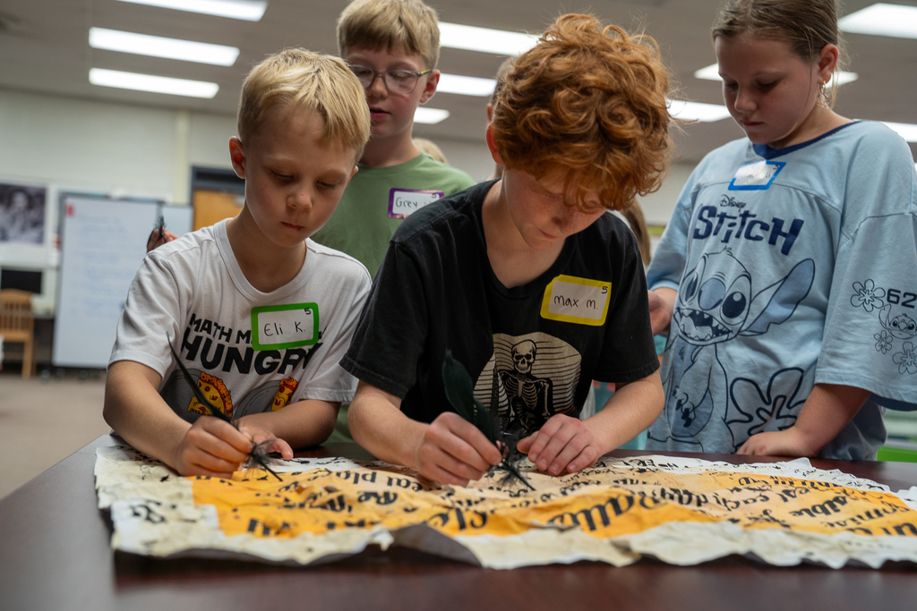 A student with blonde hair and a student with red curly hair writing on a crumbled parchment with inked quills
