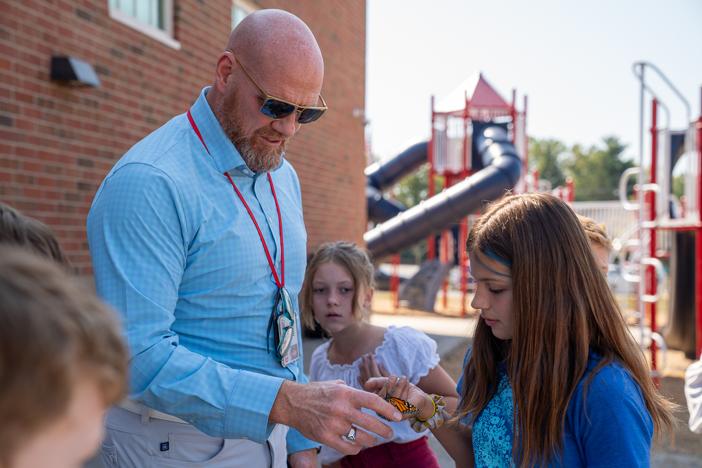 A bald man with sunglasses holding out his hand with a girl placing a butterfly on his finger