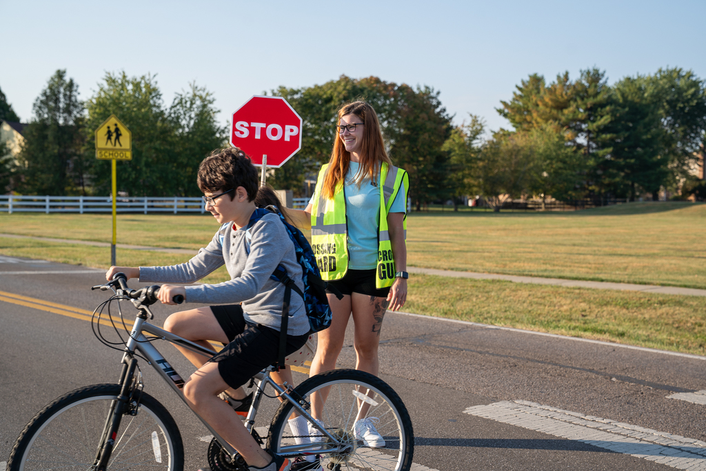 Woman smiling while holding a stop sign while a boy on a bicycle strolls by her