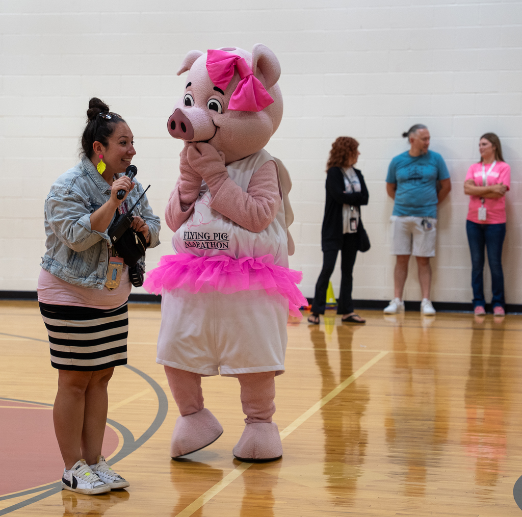a pig mascot speaking with a woman in a denim jacket in a gym