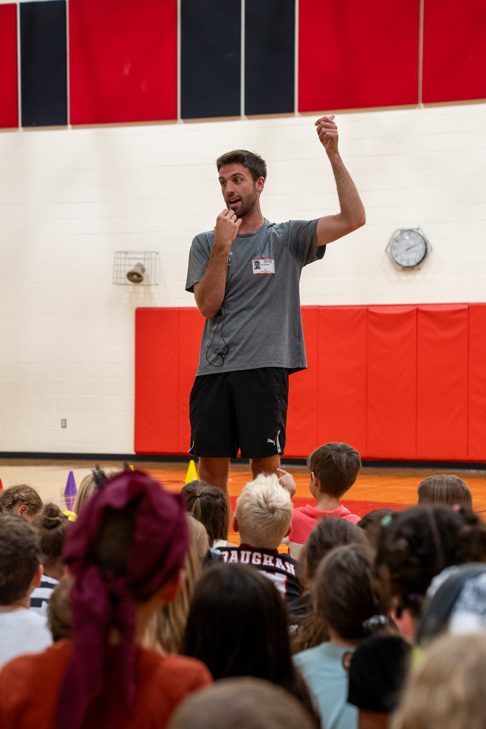 a man with a grey shirt and microphone talking to a gym full of children