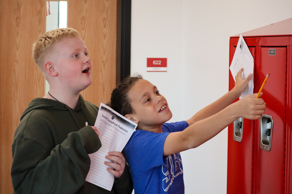 Two kids next  to red lockers with paper and pencil pointing upwards 