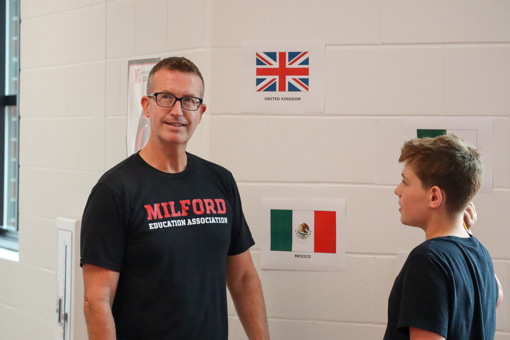 A man in a Milford shirt smiling upwards with pictures of flags behind him 