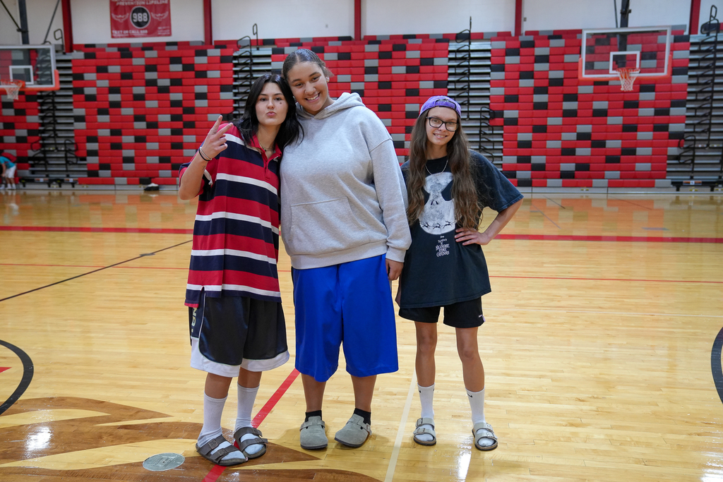 Three students standing in a gymnasium. (Left to Right) girl wearing a polo shirt and gym shorts, gurk wearing grey sweatshirt and blue gym shorts, girl wearing oversized tee shirt and backwards hat