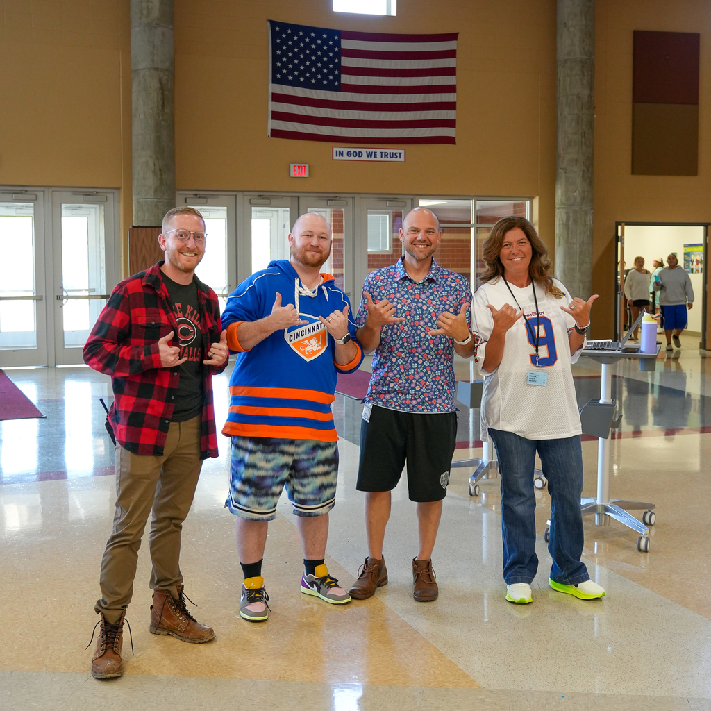 Group of adults (Left to Right) man with red flannel next to man in oversized FC Cincinnati sweatshirt next to man in hawaiian shirt and gym shorts, next to woman wearing white 9 jersey 