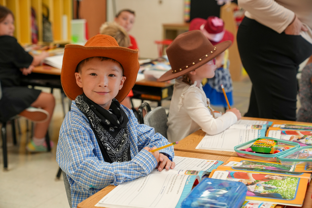 A young boy in a button down shirt wearing a cowboy hat smiling at the camera while reading