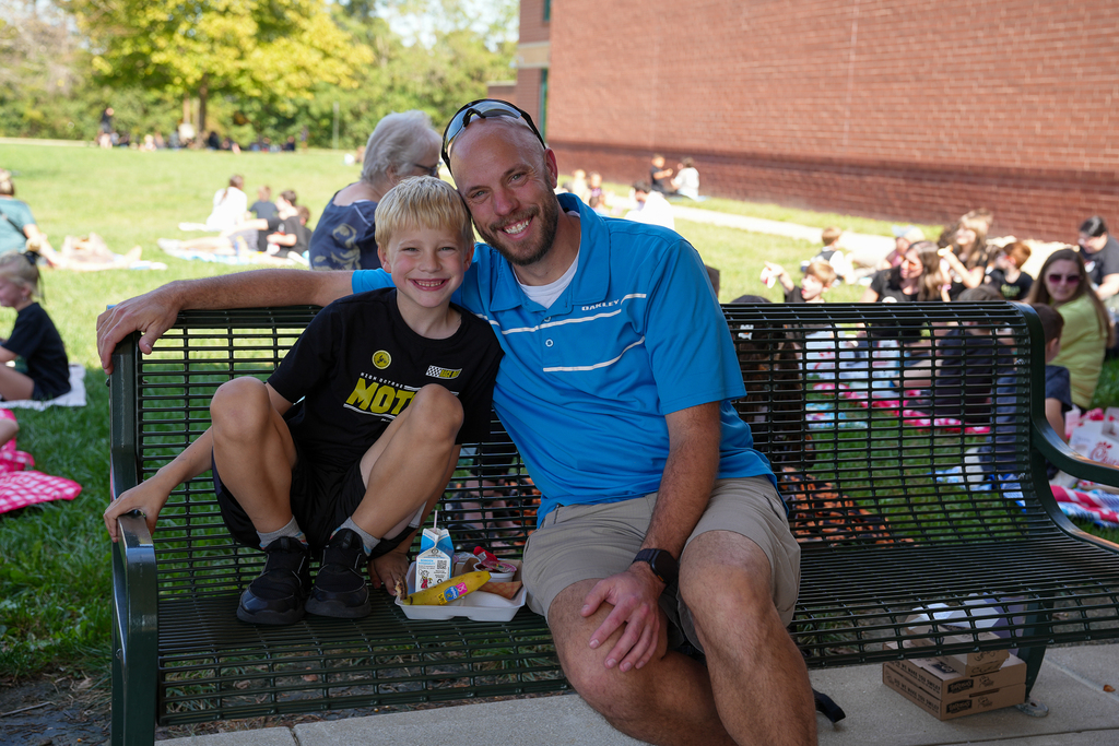 A bald man sitting with a boy with blonde hair and glasses on a bench