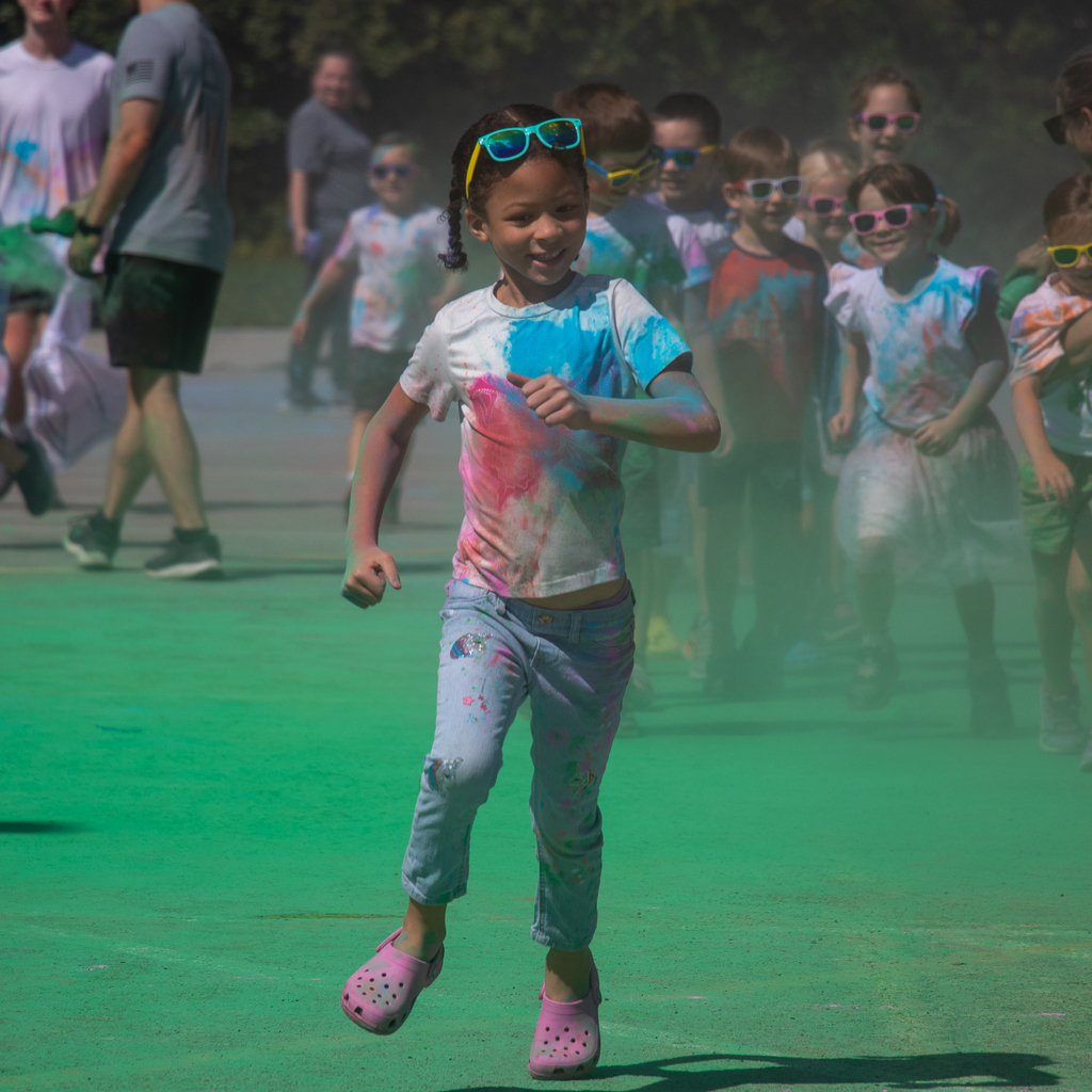 a girl covered in colorful powders jogging and smiling in a green covered sidewalk