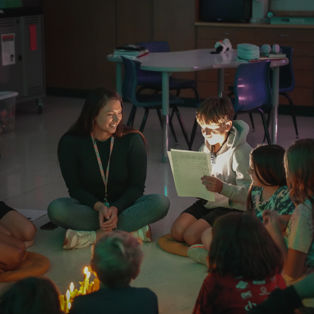 A teacher wearing jeans in a dark room next to a boy reading a piece of paper