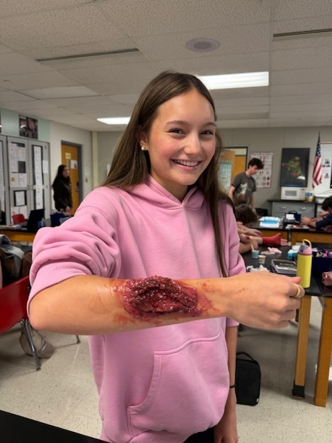 A girl in a pink sweatshirt holding out her arm covered in a fake flesh wound