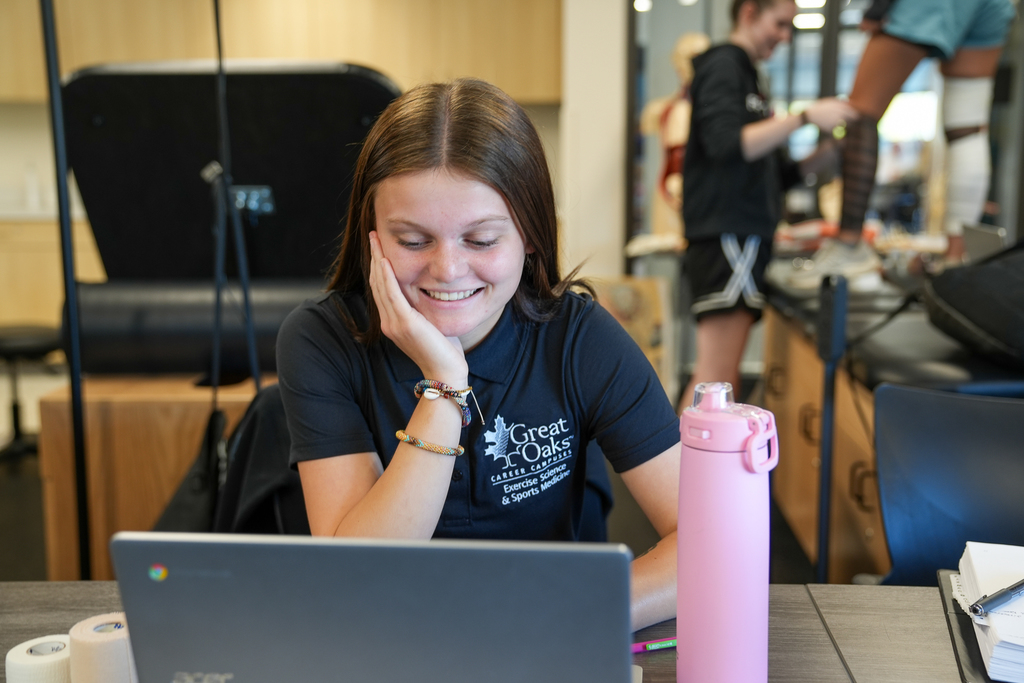 A girl with a "Great Oaks" shirt smiling at her laptop