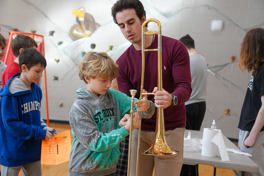 A student with blonde hair holding a trombone with another male helping