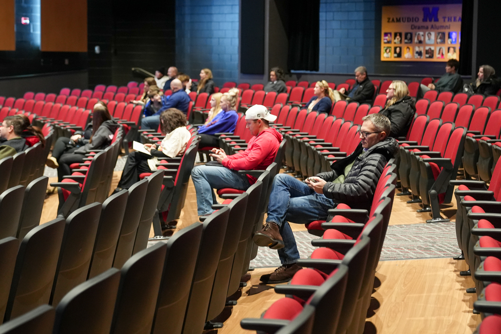 Parents and students sit spaced throughout a school auditorium with red seats, listening and looking at their phones or papers during a schedule fair presentation.