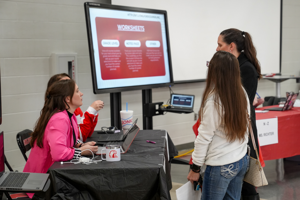 Staff members sit at a table with laptops and a large screen labeled “Worksheets,” assisting parents and students with course selection questions.