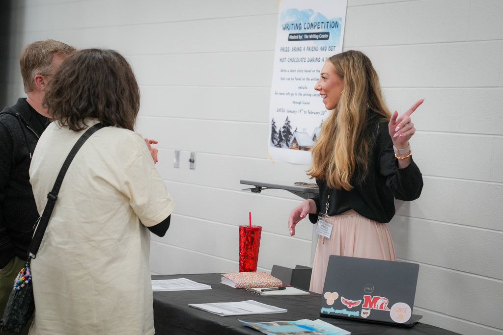 A teacher stands behind a table with a laptop and papers, pointing while speaking with parents near a poster about a writing competition at a high school schedule fair.