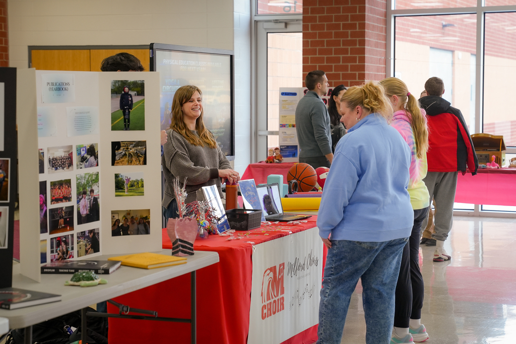 A teacher smiles while speaking with parents at a red-covered table featuring a publications display board and choir information during a high school schedule fair.