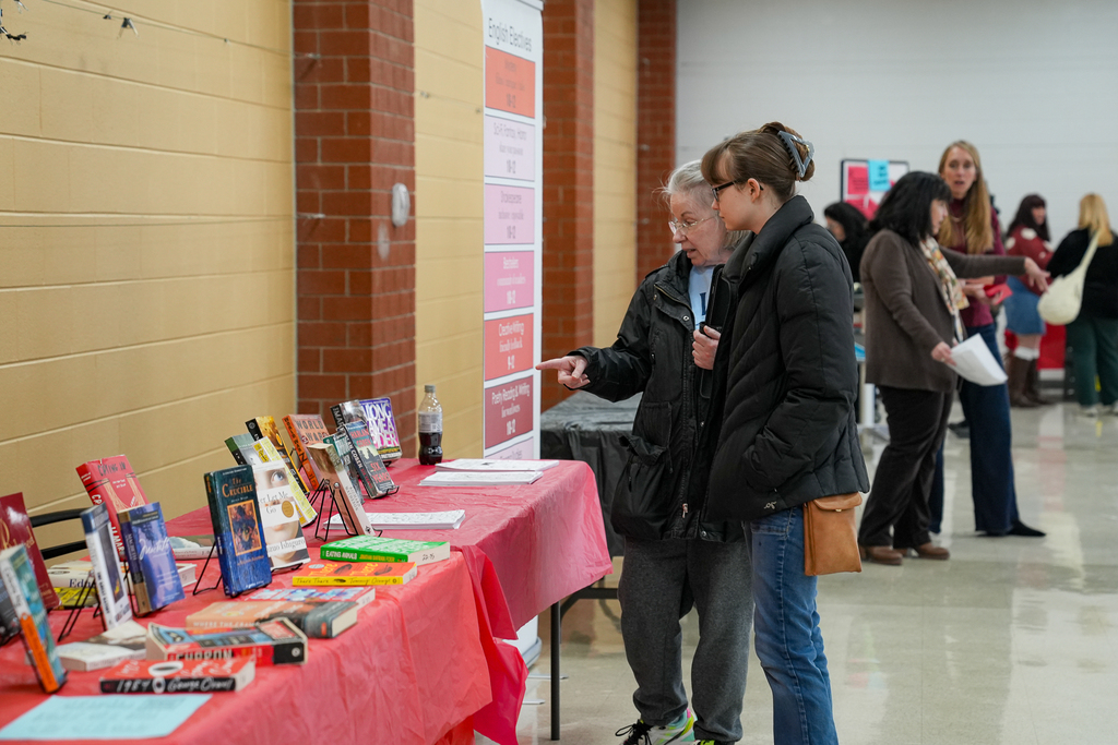 Two adults stand at a table covered in red cloth, looking at a display of books and course materials set up along a school hallway during a schedule fair.