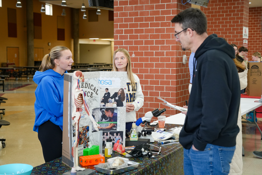 Two students present a HOSA medical science display with a model skeleton and microscope while speaking with a parent at a table during a school course fair.