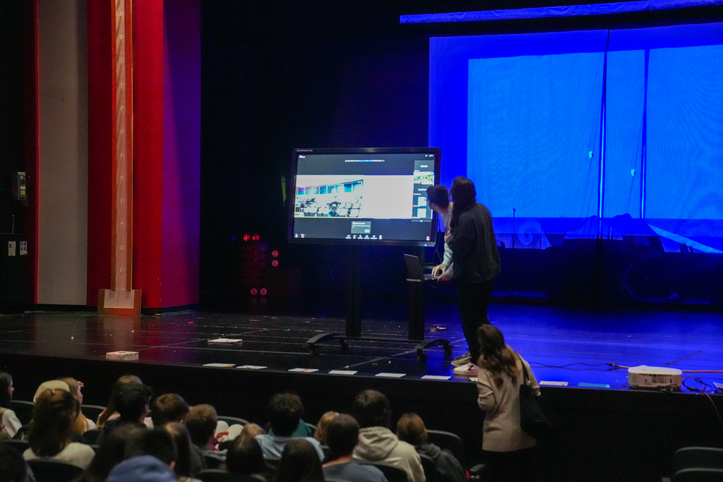 Two presenters stand beside a large digital screen on a stage while students watch from the auditorium during a live-streamed brain surgery seminar.