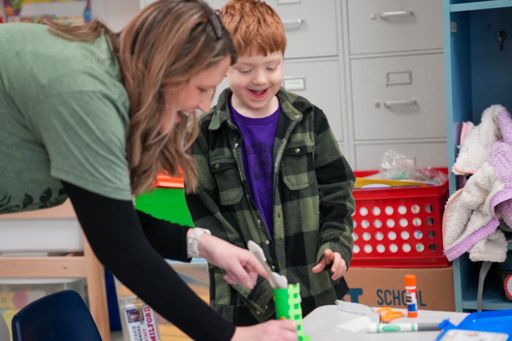 A teacher leans over a table, smiling as she helps a young student build a green leprechaun trap using classroom craft materials. The student, wearing a green plaid shirt, looks excited while working on the project. Classroom supplies and bins are visible in the background.