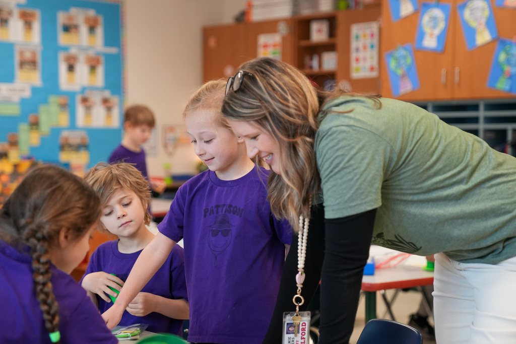 A teacher leans over a classroom table, smiling as she assists a small group of young students wearing purple shirts. The students focus on assembling materials for a leprechaun trap, with colorful classroom displays and cabinets visible in the background.