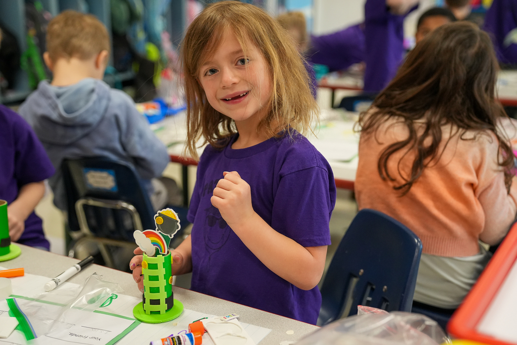 A young student in a purple shirt smiles at the camera while holding a handmade leprechaun trap decorated with a rainbow and pot of gold. Other students work on similar crafts at tables in the background, surrounded by classroom supplies.