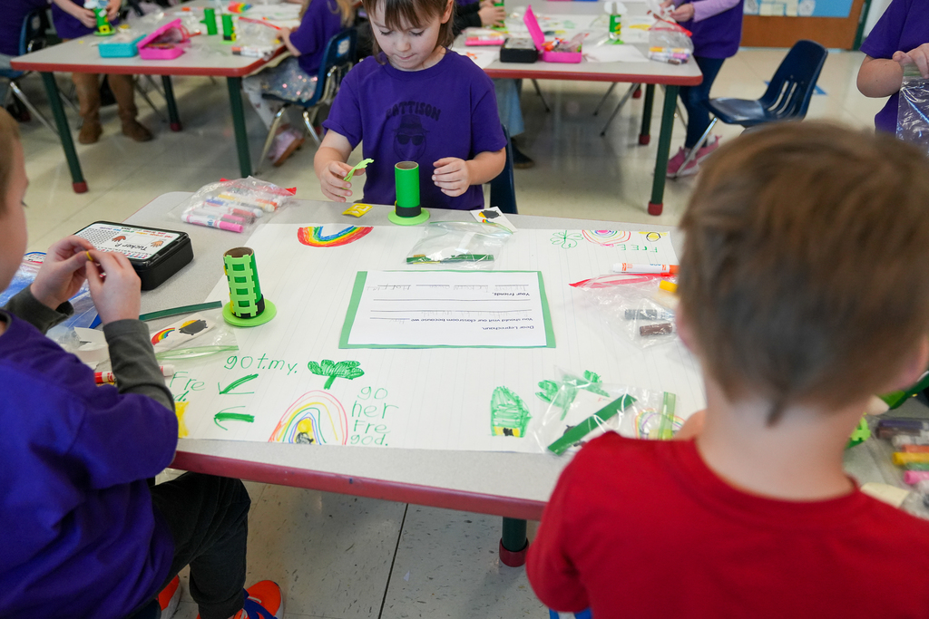 Students sit around a classroom table working on leprechaun trap crafts, using markers, paper, and small materials. One student in a purple shirt focuses on assembling a green trap while colorful drawings of rainbows and clovers are spread across the table.