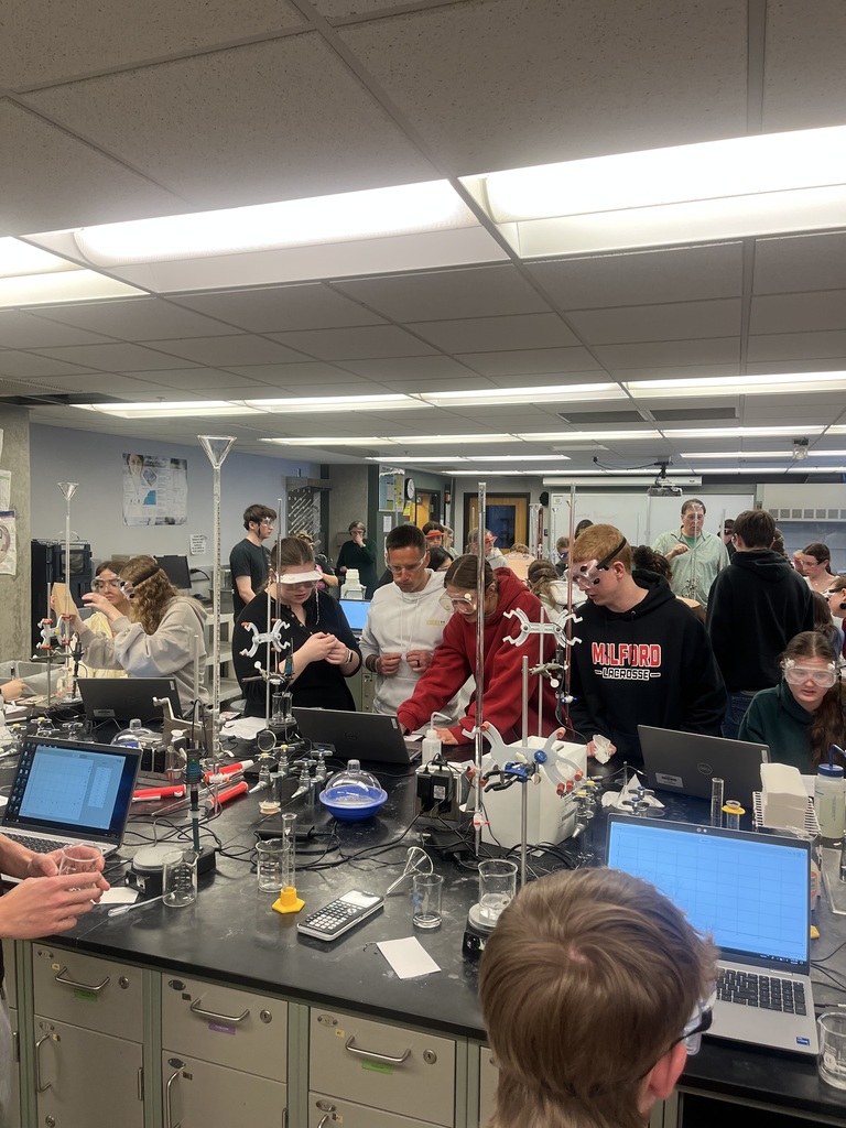 Students wearing safety goggles work together at a lab station in a college chemistry lab, using glassware and equipment while measuring and recording data.