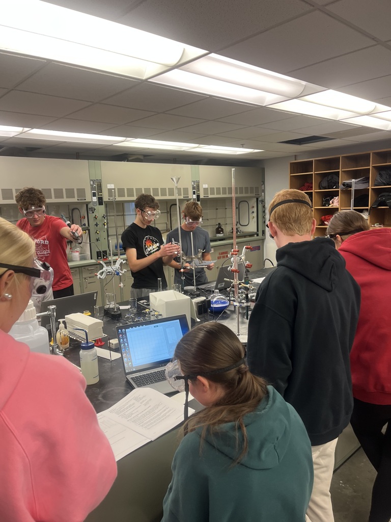 A group of students in safety goggles conduct a chemistry experiment at a lab table, handling equipment and following instructions on a worksheet and laptop.