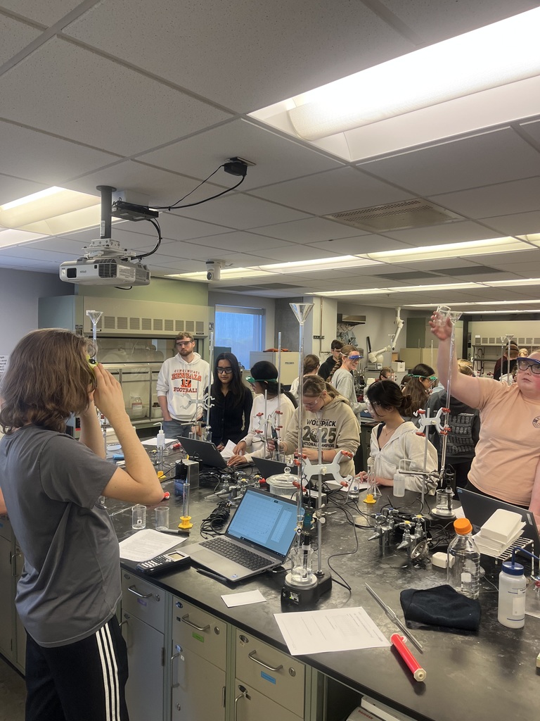 Students wearing safety goggles work together at a lab station in a college chemistry lab, using glassware and equipment while measuring and recording data.