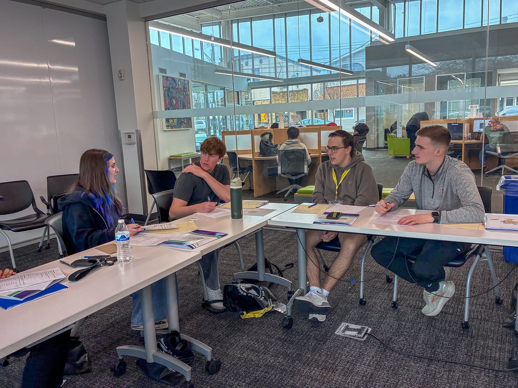 A small group of students sits around tables in a modern classroom, discussing and writing on worksheets while a facilitator leads the conversation.