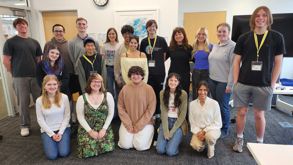 A group of students and adults pose together indoors, smiling at the camera, many wearing name badges after a collaborative or training session.