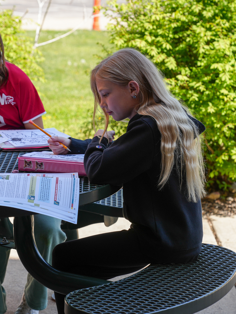 An elementary student sits at an outdoor picnic table, focused on writing in a workbook, with papers and a textbook spread out beside them on a sunny day.