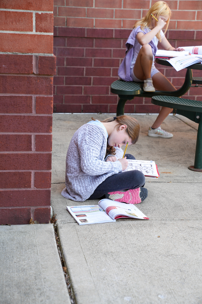 Two elementary students sit outside near a brick wall, working independently on assignments. One student sits on the ground writing in a notebook while another sits at a picnic table reading and writing.