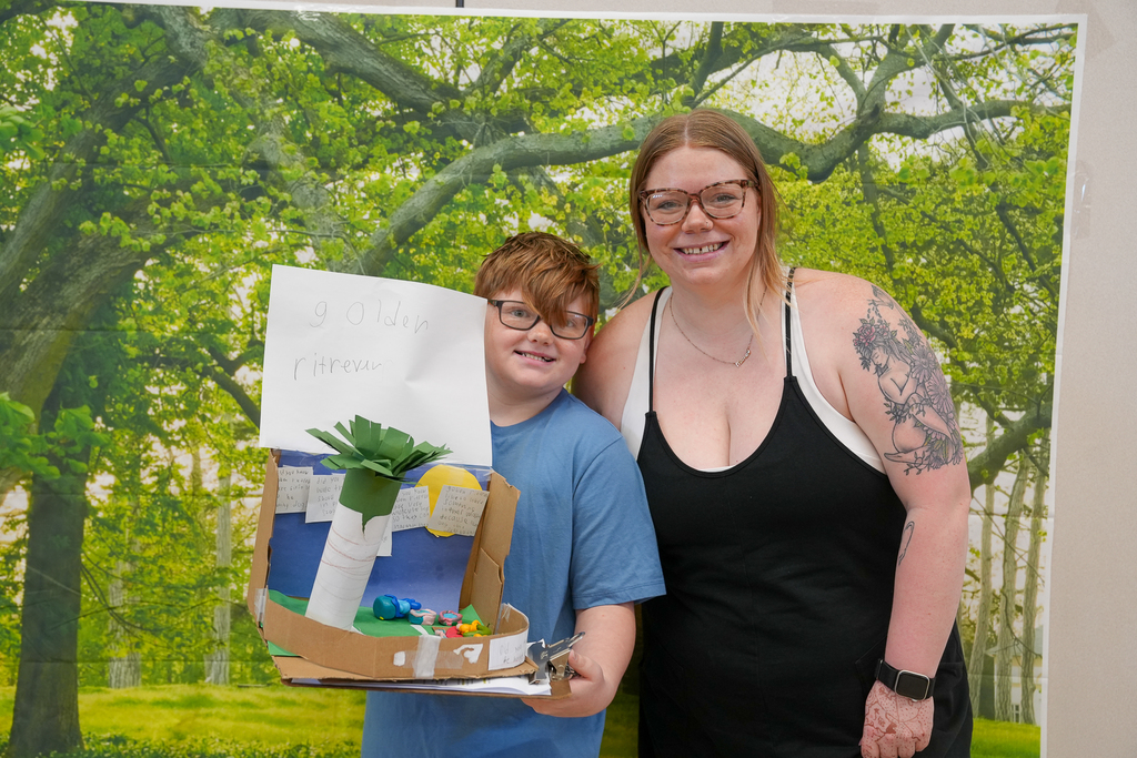 A smiling student stands next to an adult while holding a handmade diorama labeled “golden retriever,” featuring a small scene with paper and craft materials.