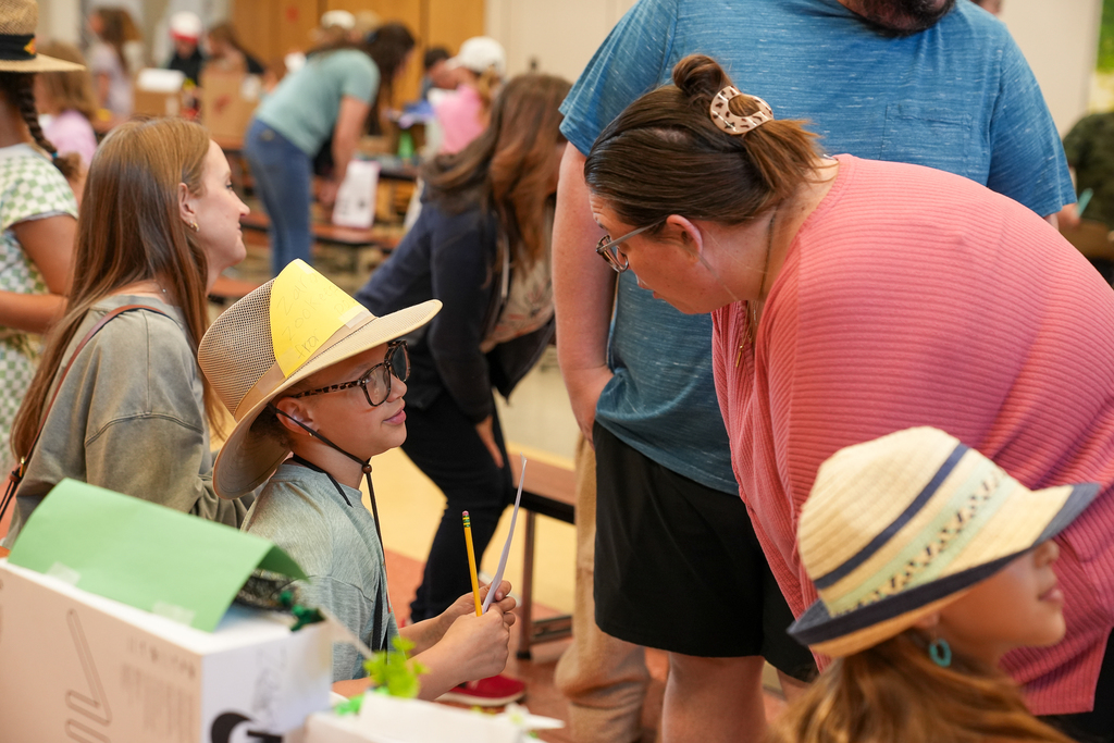 An adult leans in to talk with a student wearing a wide-brim hat and holding a clipboard while presenting a project, with others gathered around nearby displays.