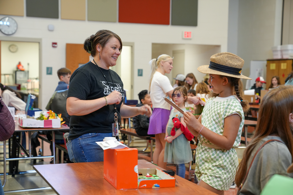 A teacher speaks with a student holding a clipboard at a table displaying a habitat project, while other students and families interact in the background.