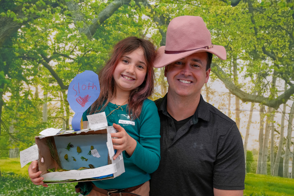 A smiling student stands next to an adult while holding a handmade “sloth” habitat project, with a forest backdrop behind them.